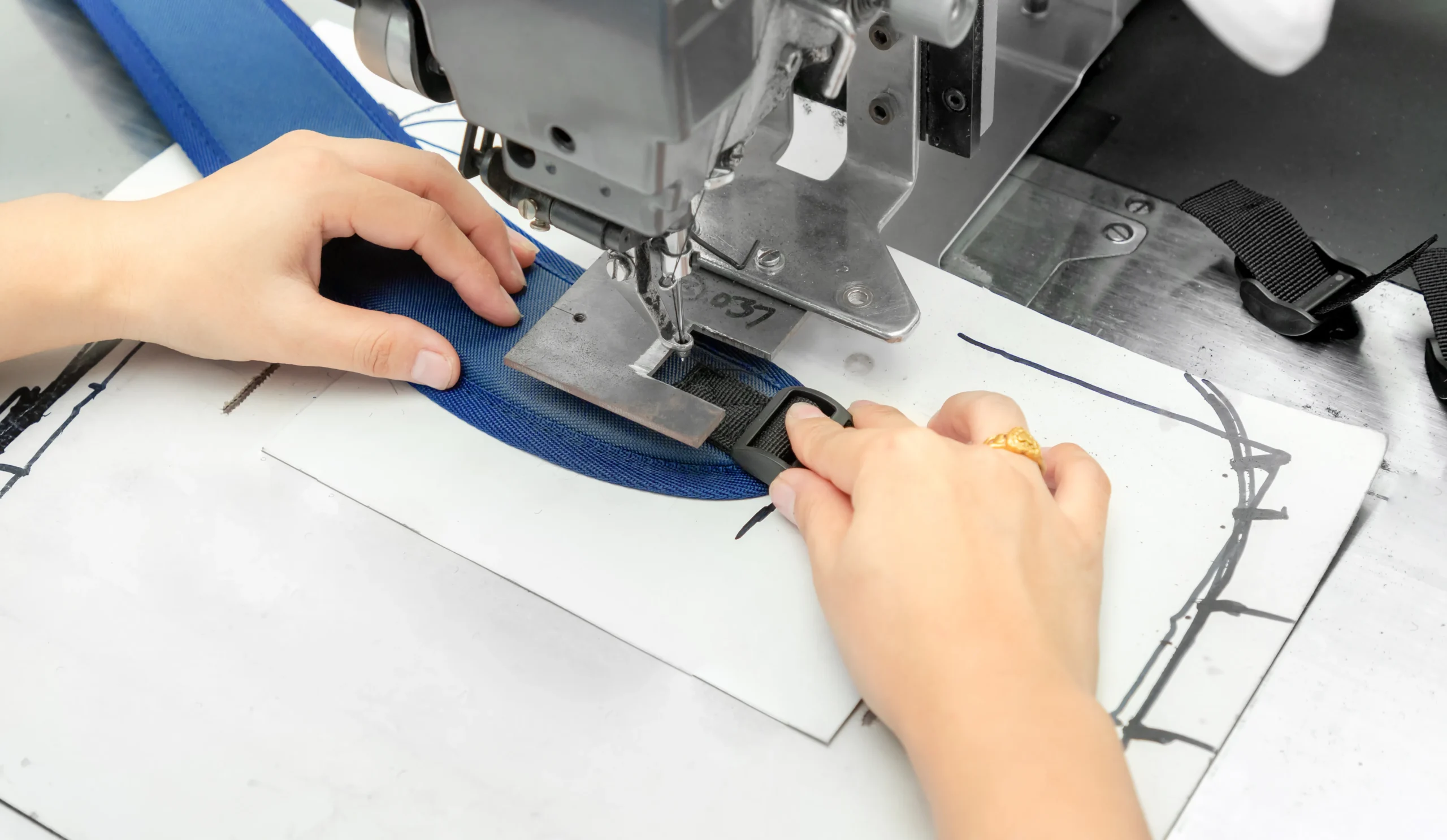 Close-up of sewing a blue strap and plastic buckle onto a backpack. OEM Supplier production detail.