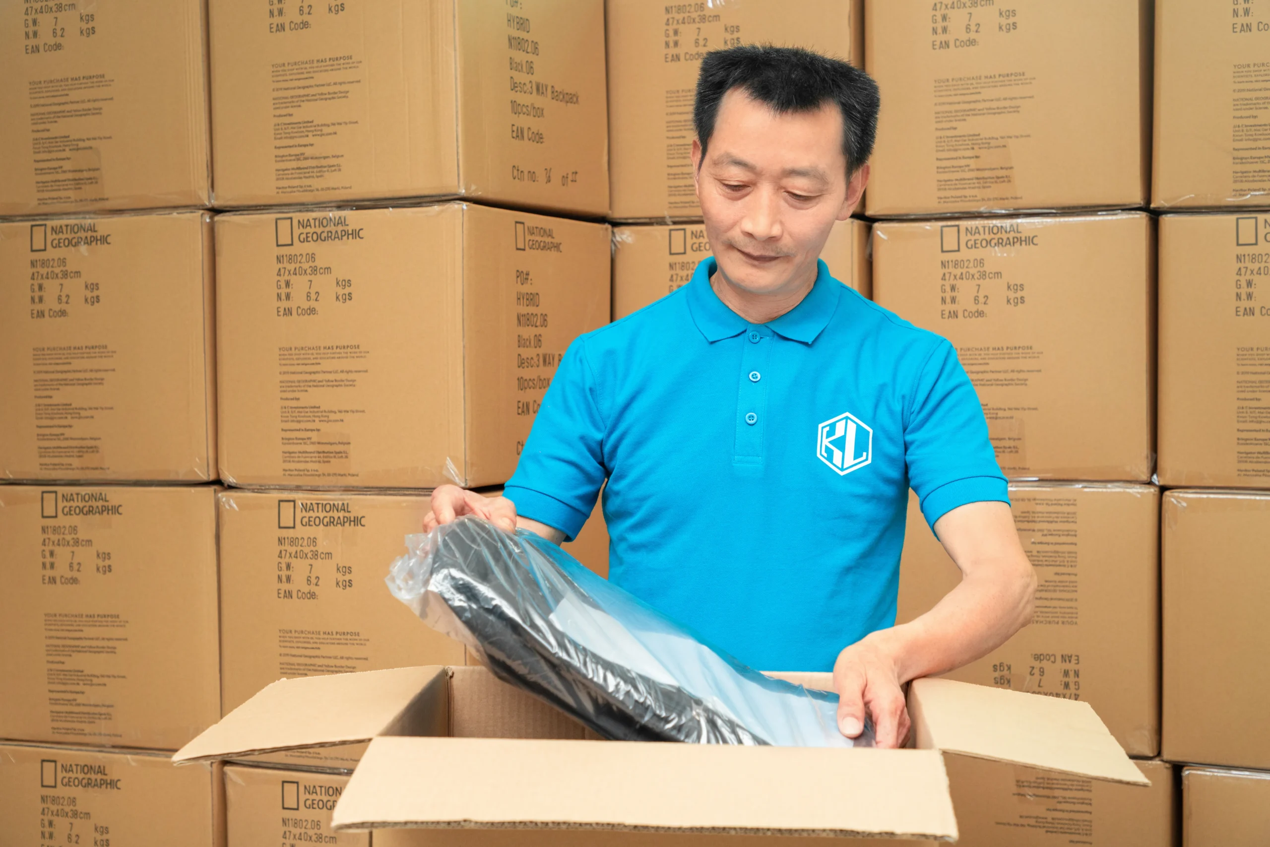 Male worker removing a packaged backpack from a National Geographic box. Bulk packaging and inspection by a Backpack Manufacturer.