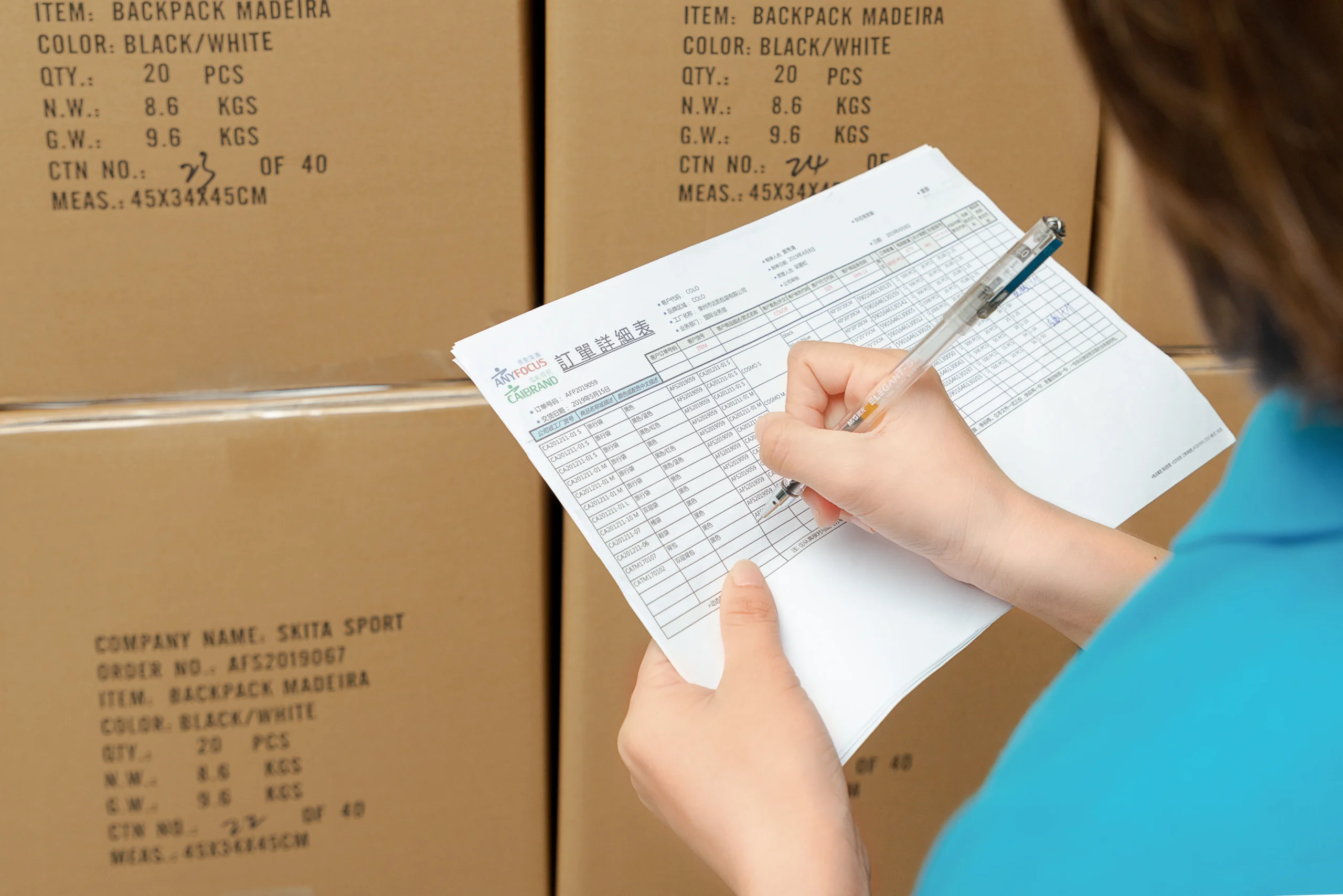 Worker reviewing a detailed packing list against shipping cartons. Logistics and inventory control by a Backpack Manufacturer.