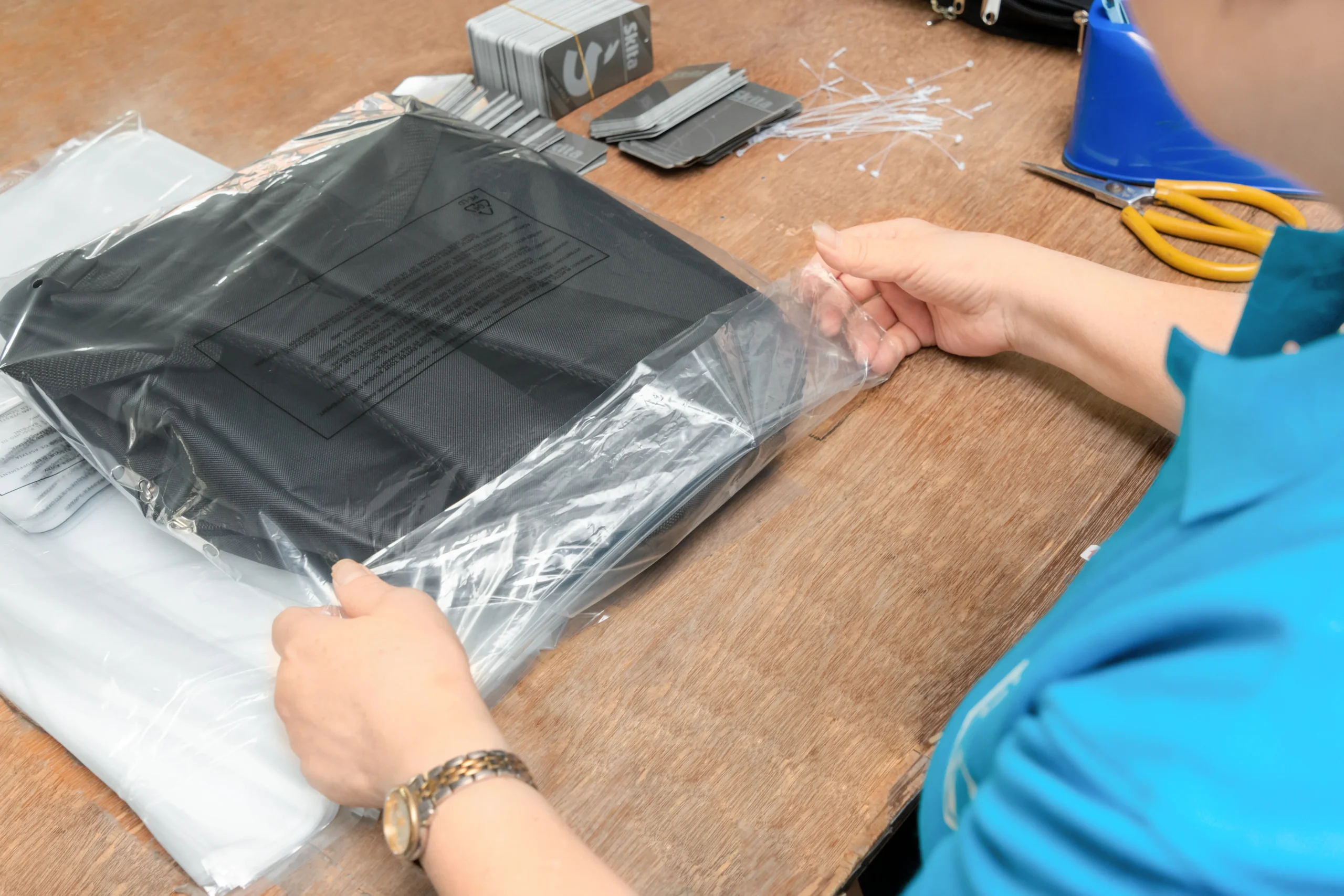 Hands wrapping a black backpack in clear plastic. Protective packaging at a Backpack Manufacturer and Supplier.