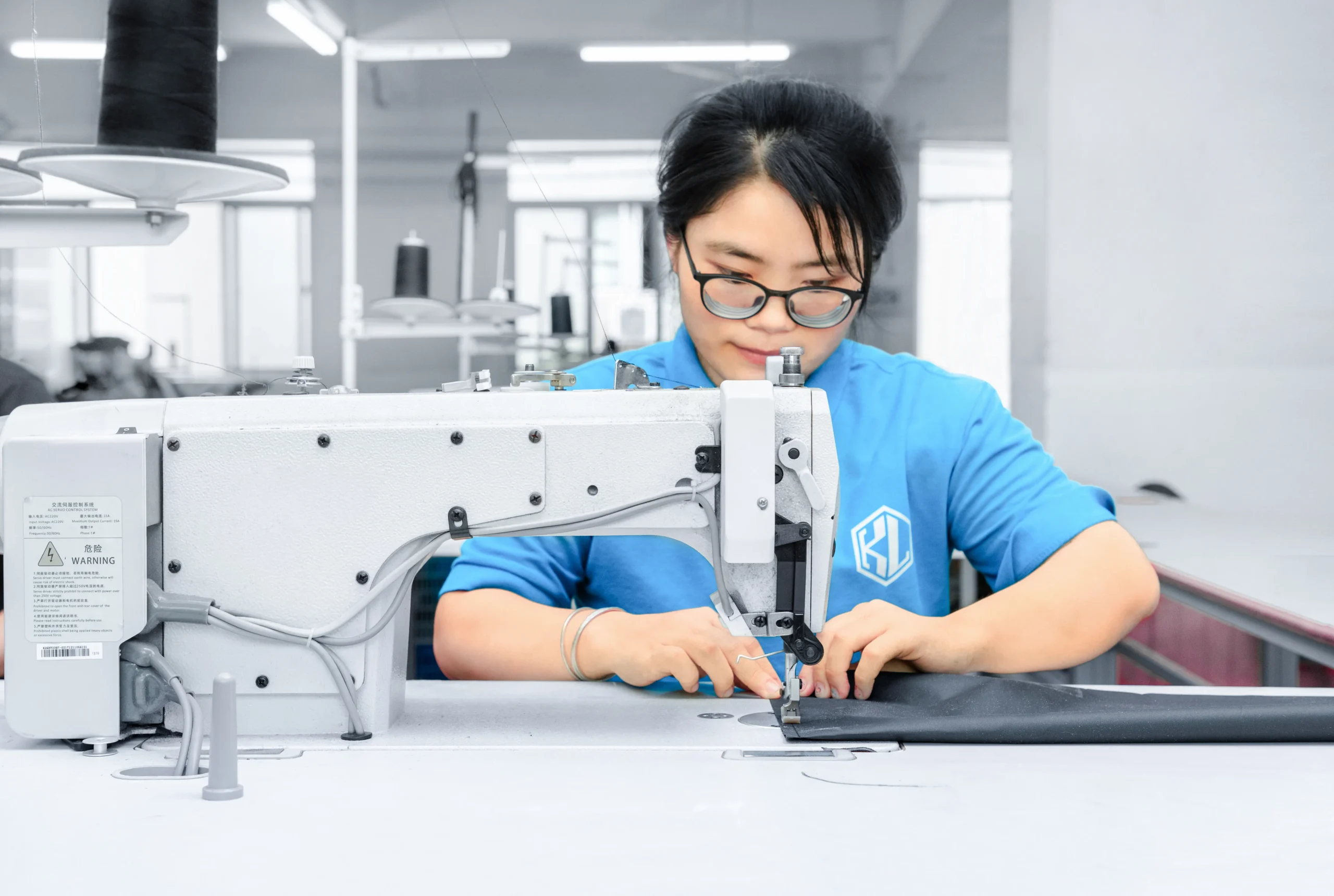 Female worker wearing glasses operating a white sewing machine. Skillful sewing staff at a custom Backpack Manufacturer.