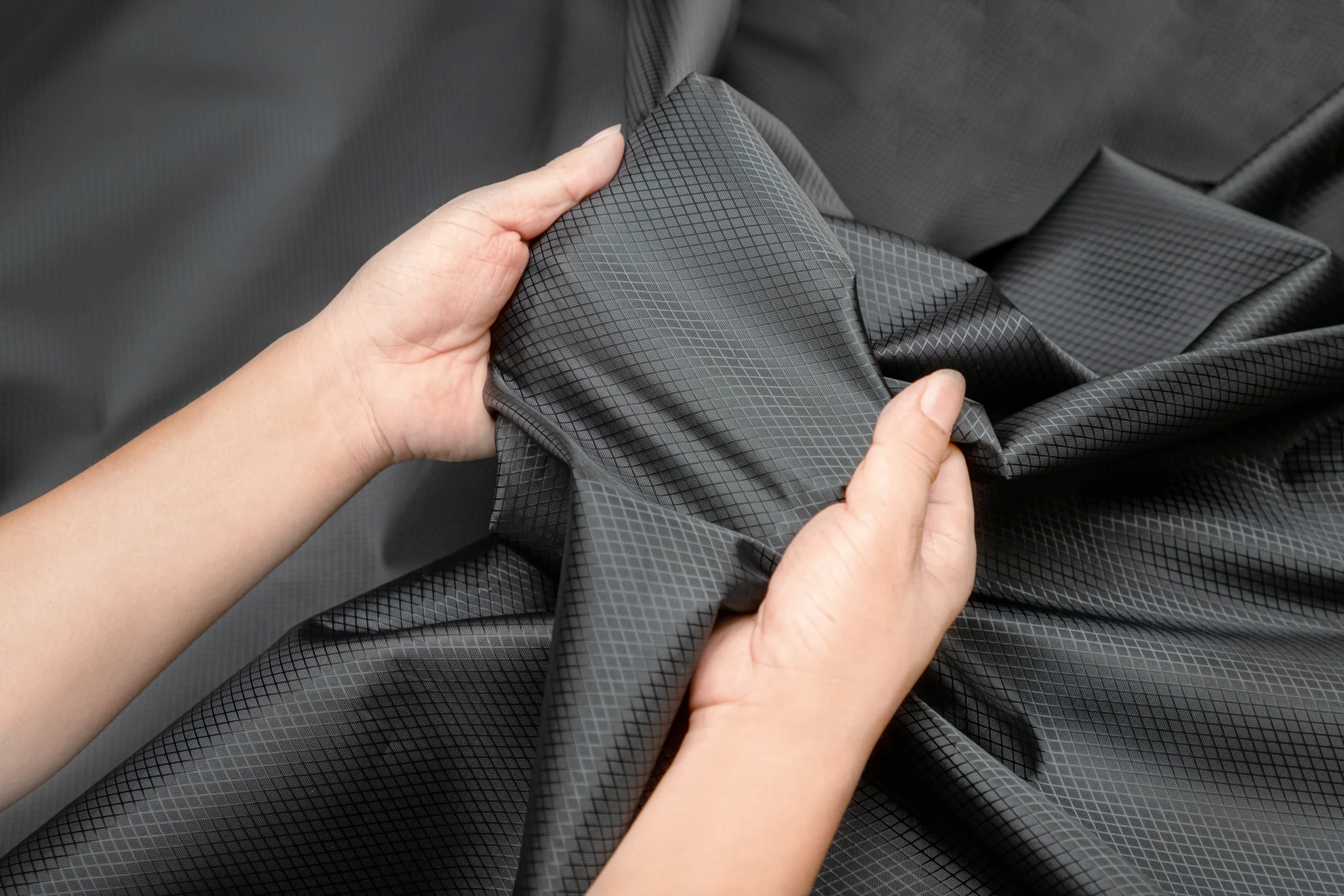 Hands testing the stretch of black ripstop fabric. Physical material testing at a Backpack OEM Supplier.