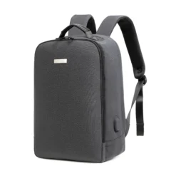 Slanted front view of a minimalist dark grey business backpack with a sleek silver logo plate and a side charging port. Functional smart backpack by a Custom Backpack Manufacturer.
