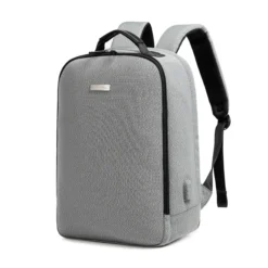 Slanted front view of a minimalist light grey business backpack with a sleek silver logo plate and a side charging port. Functional smart backpack by a Custom Backpack Manufacturer.